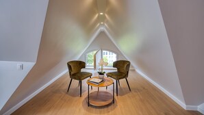 A room with a sloped ceiling, two armchairs, a round table with a book and a potted plant, and a large window with a view of buildings. - Golden Tulip De' Medici Hotel (Bruges)