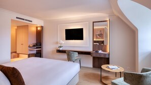 A modern hotel room with a large bed, a desk with a flat-screen TV, a chair, a small round table, and a mirror. - Golden Tulip De' Medici Hotel (Bruges)