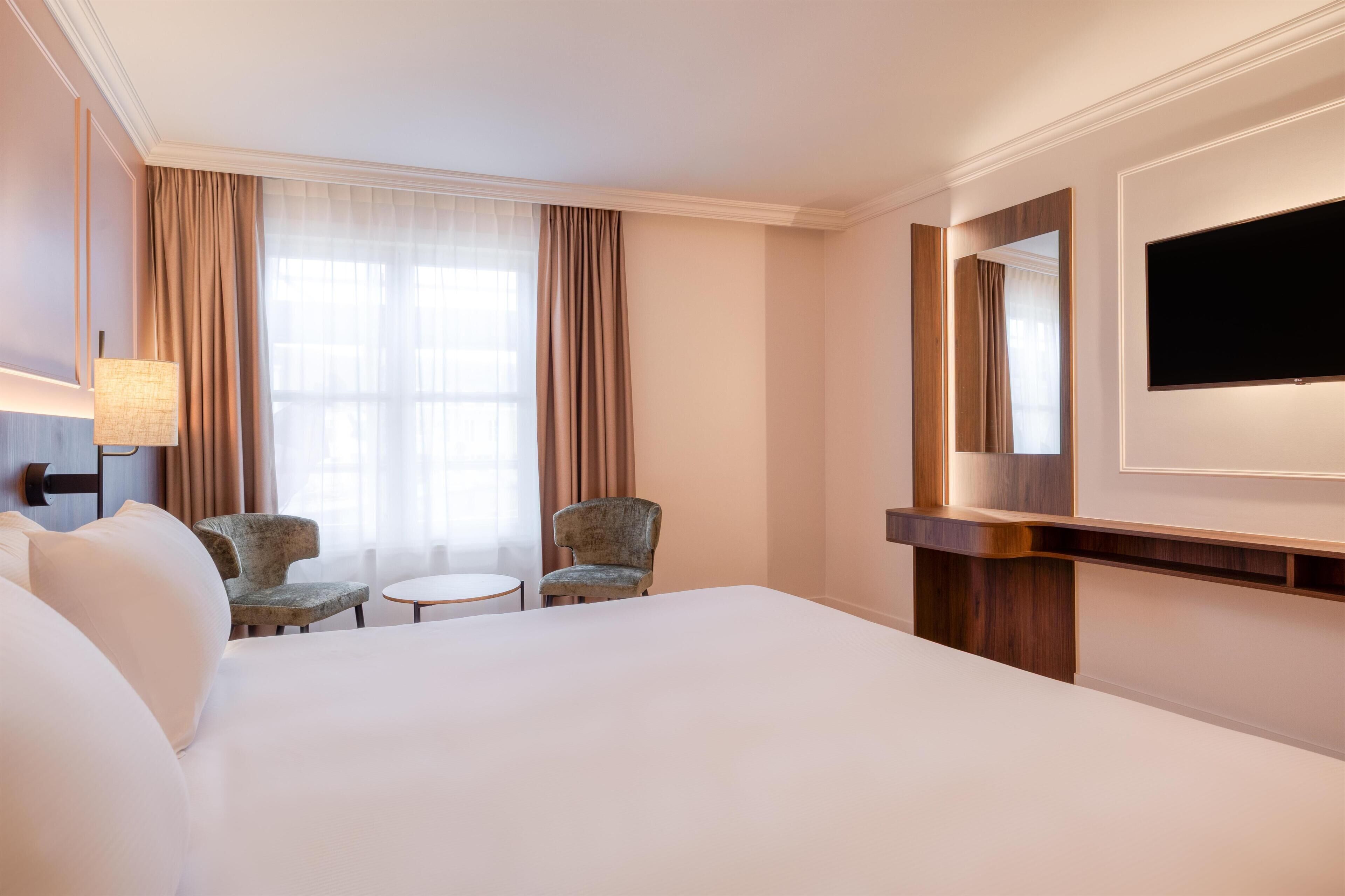 A hotel room with a large bed, two armchairs, a small round table, a TV mounted on the wall, and a large window with curtains.