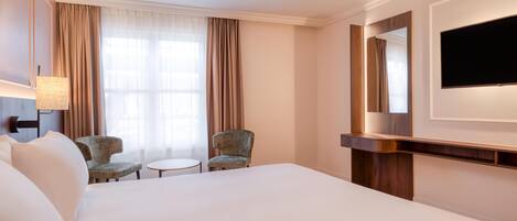 A hotel room with a large bed, two armchairs, a small round table, a TV mounted on the wall, and a large window with curtains.