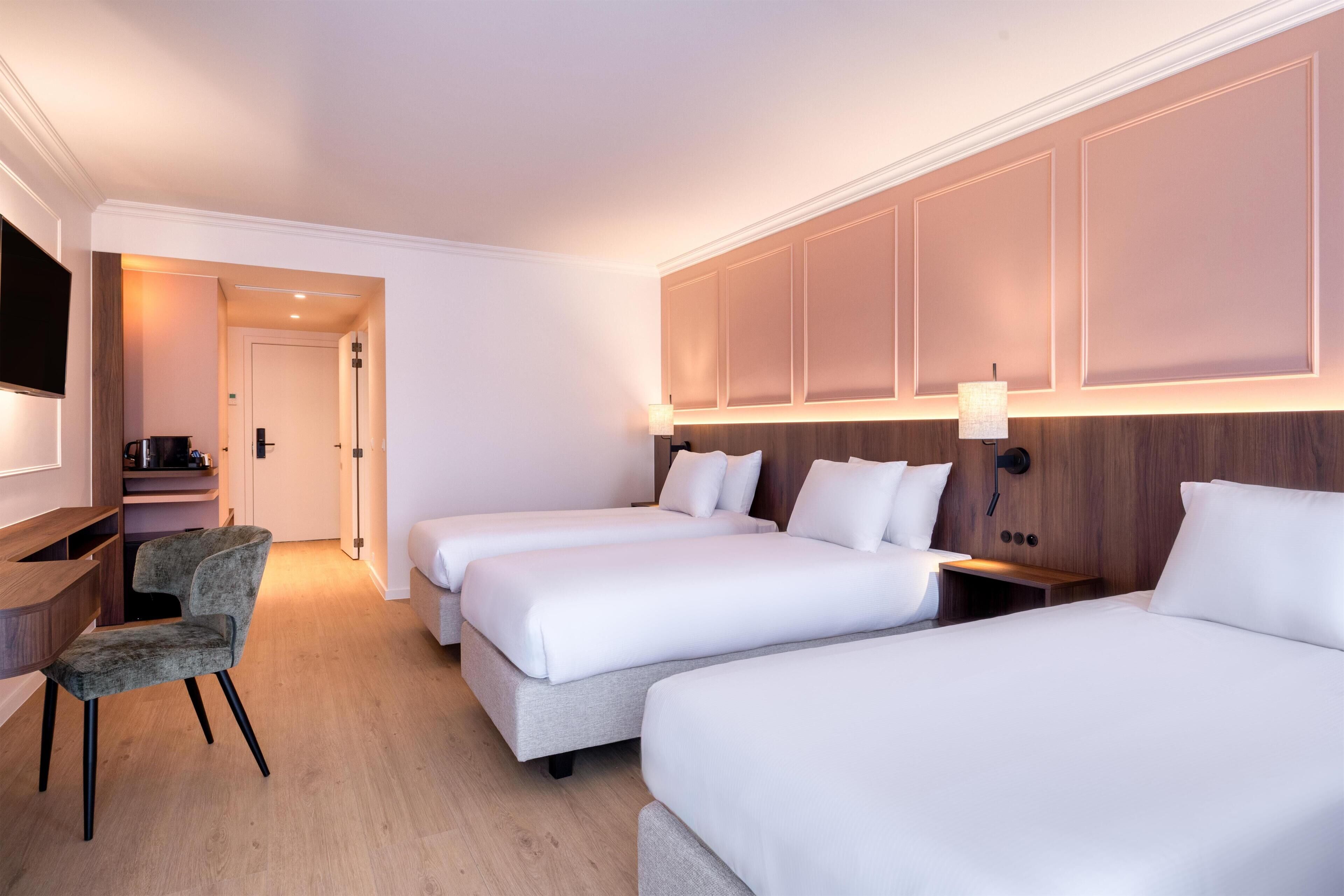 A hotel room with three beds, a desk, a chair, and a TV.