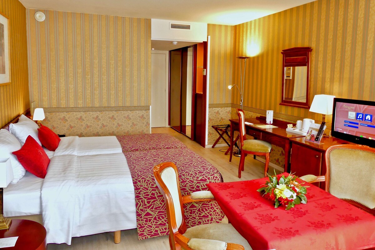 Habitación de hotel con dos camas, un escritorio con computadora, una silla y una pequeña mesa con un arreglo floral.