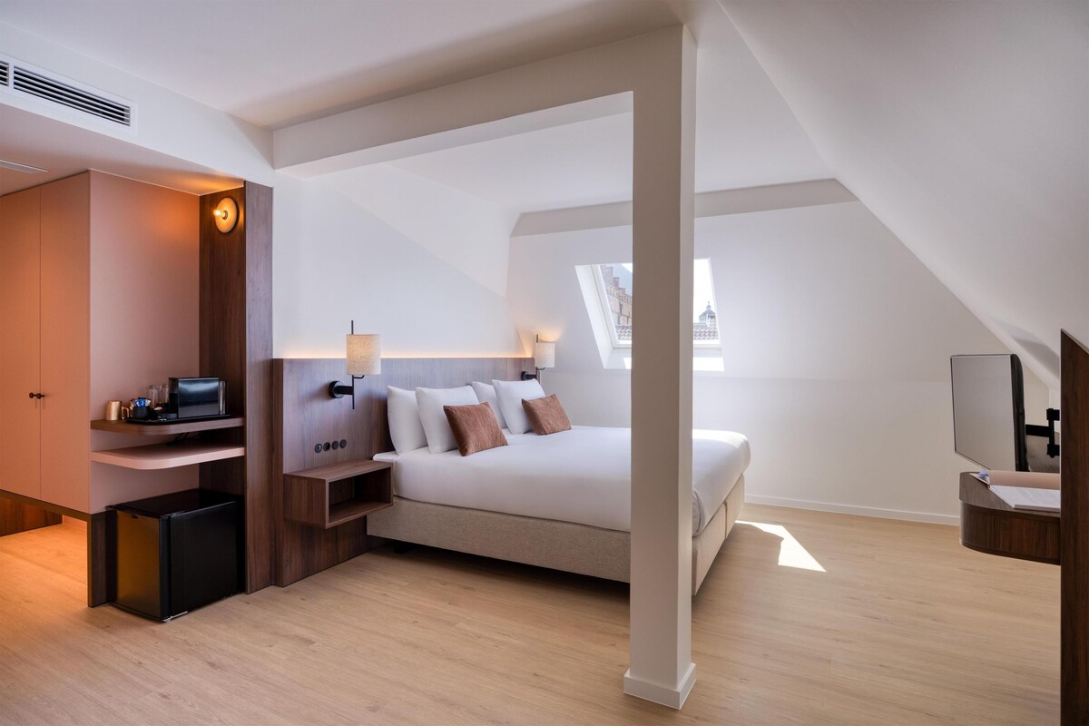 A modern bedroom with a loft-style ceiling, a bed, a desk, and a small refrigerator.