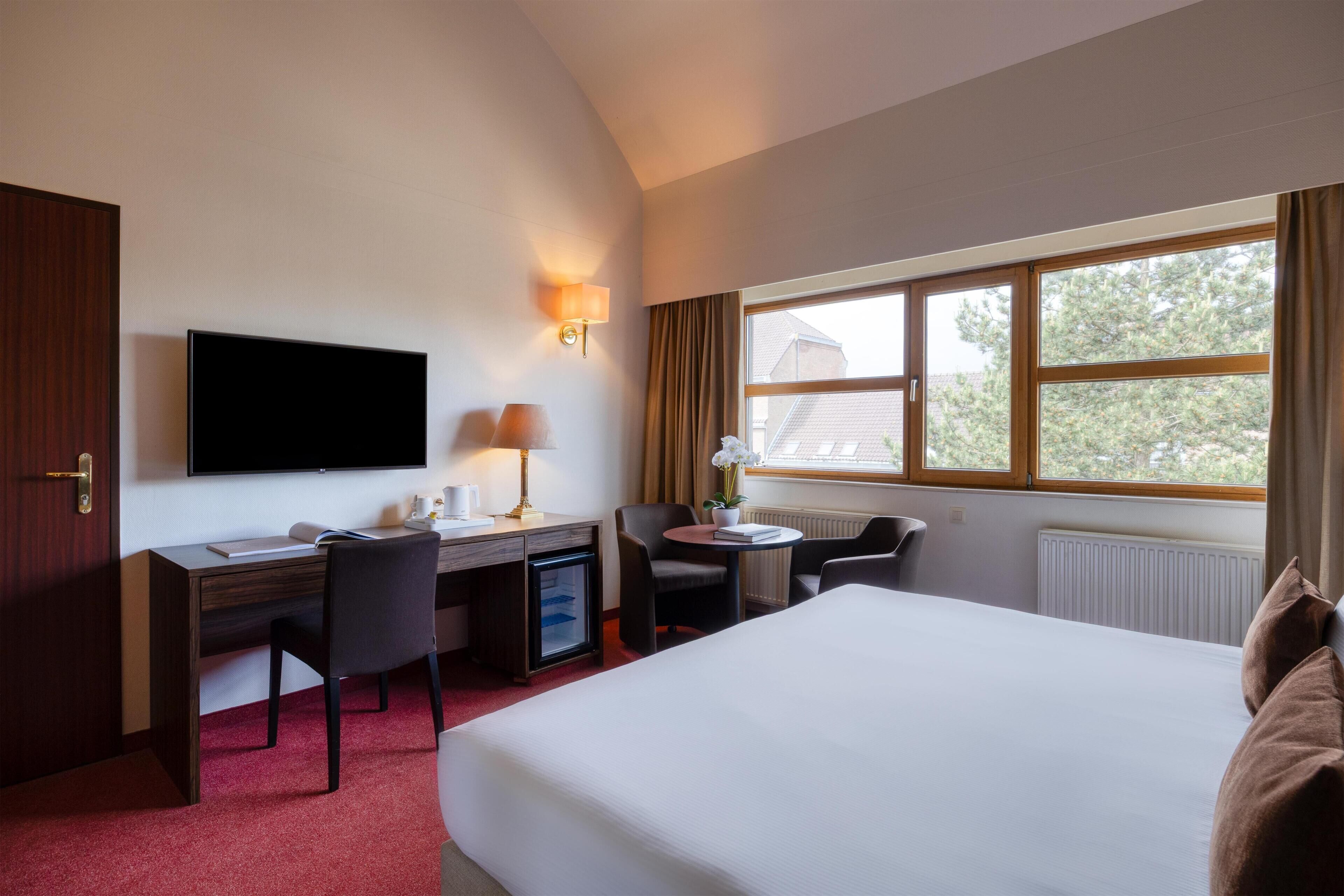 Habitación de hotel con cama, escritorio, silla, televisor y ventana con cortinas.