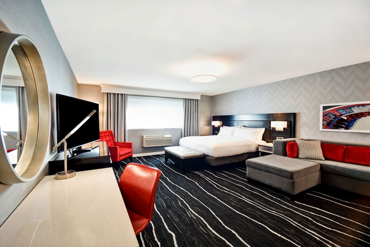 A hotel room with a bed, a desk, a TV, a red chair, a grey sofa, and a large mirror.
