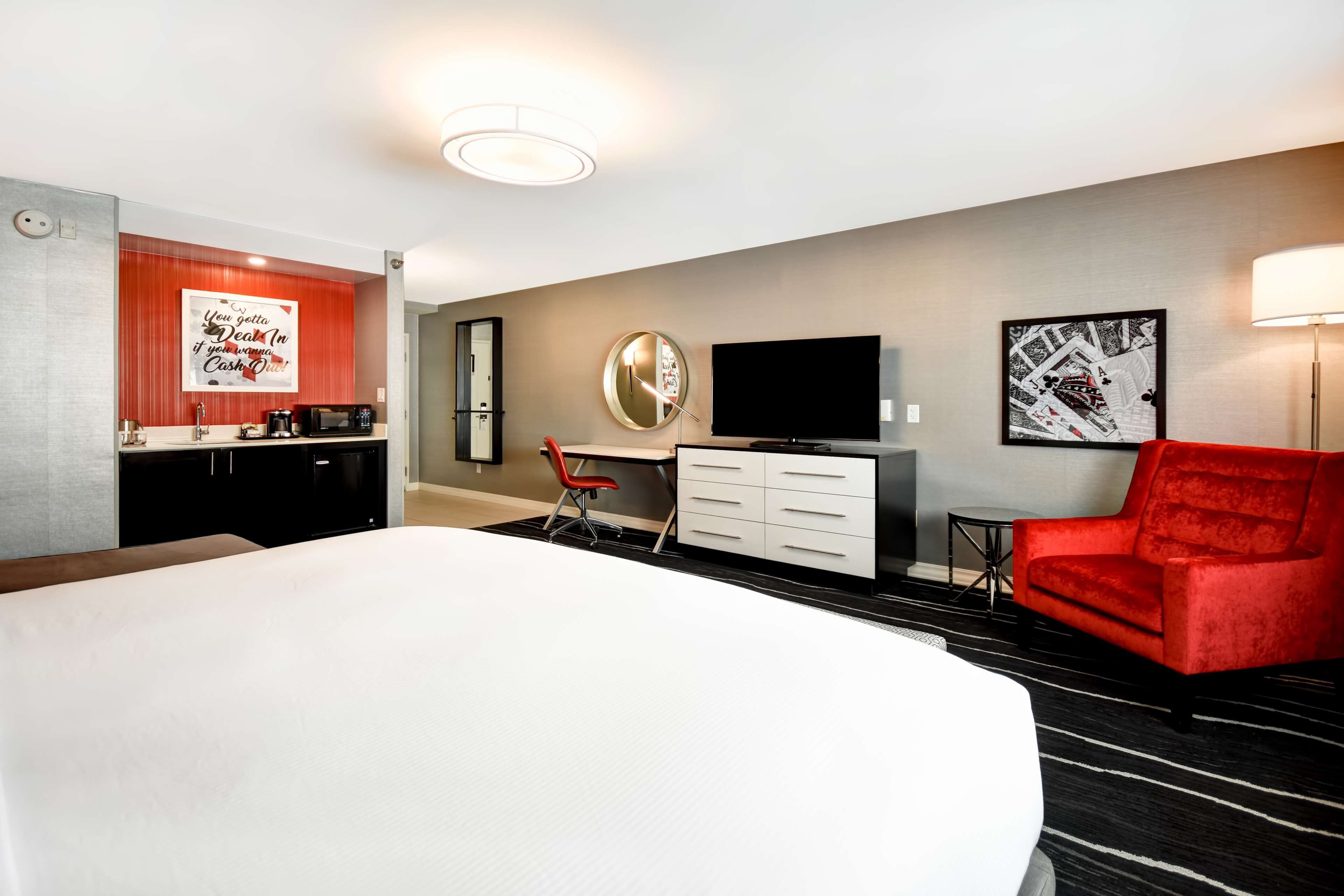 Quarto de hotel com uma cama grande, uma mesa de trabalho com cadeira, televisão e obras de arte nas paredes.