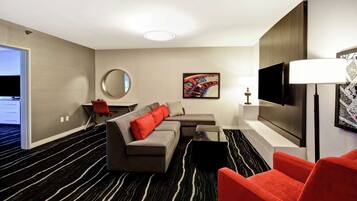 A modern hotel room with a sectional sofa, a red armchair, a flat-screen TV, and a large mirror.