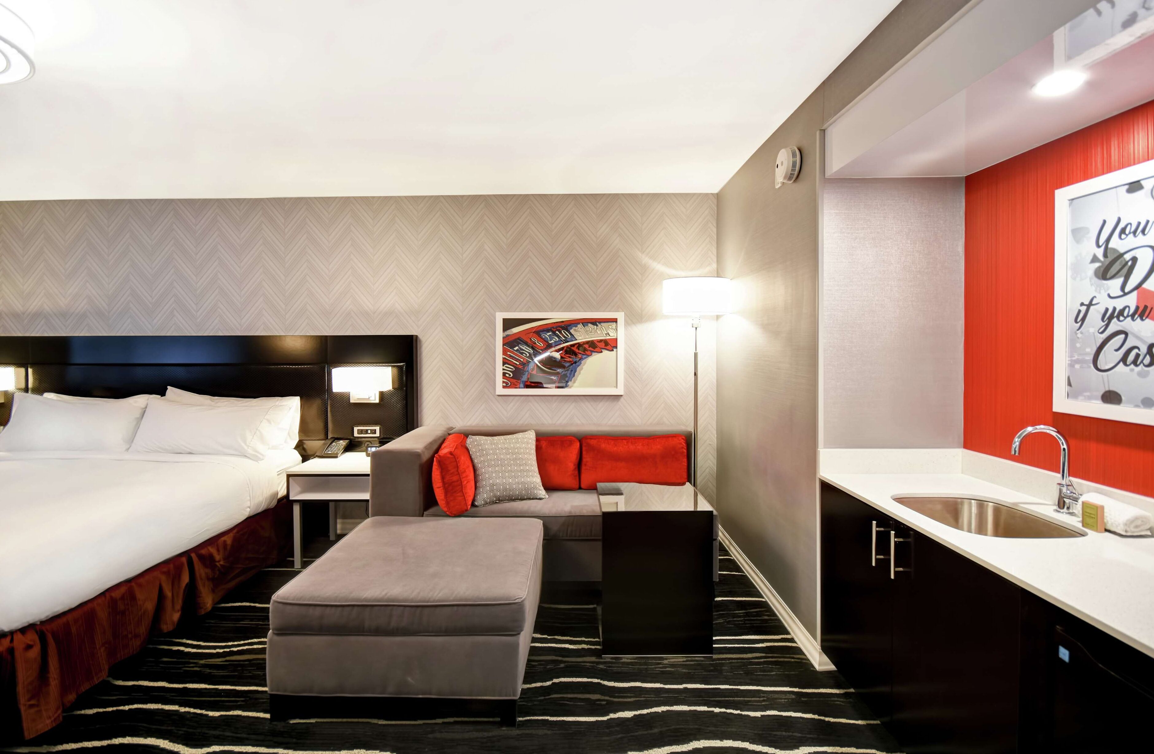 Habitación de hotel con cama, sofá, pequeña mesa, escritorio, lámpara, cartel, lavabo y espejo.