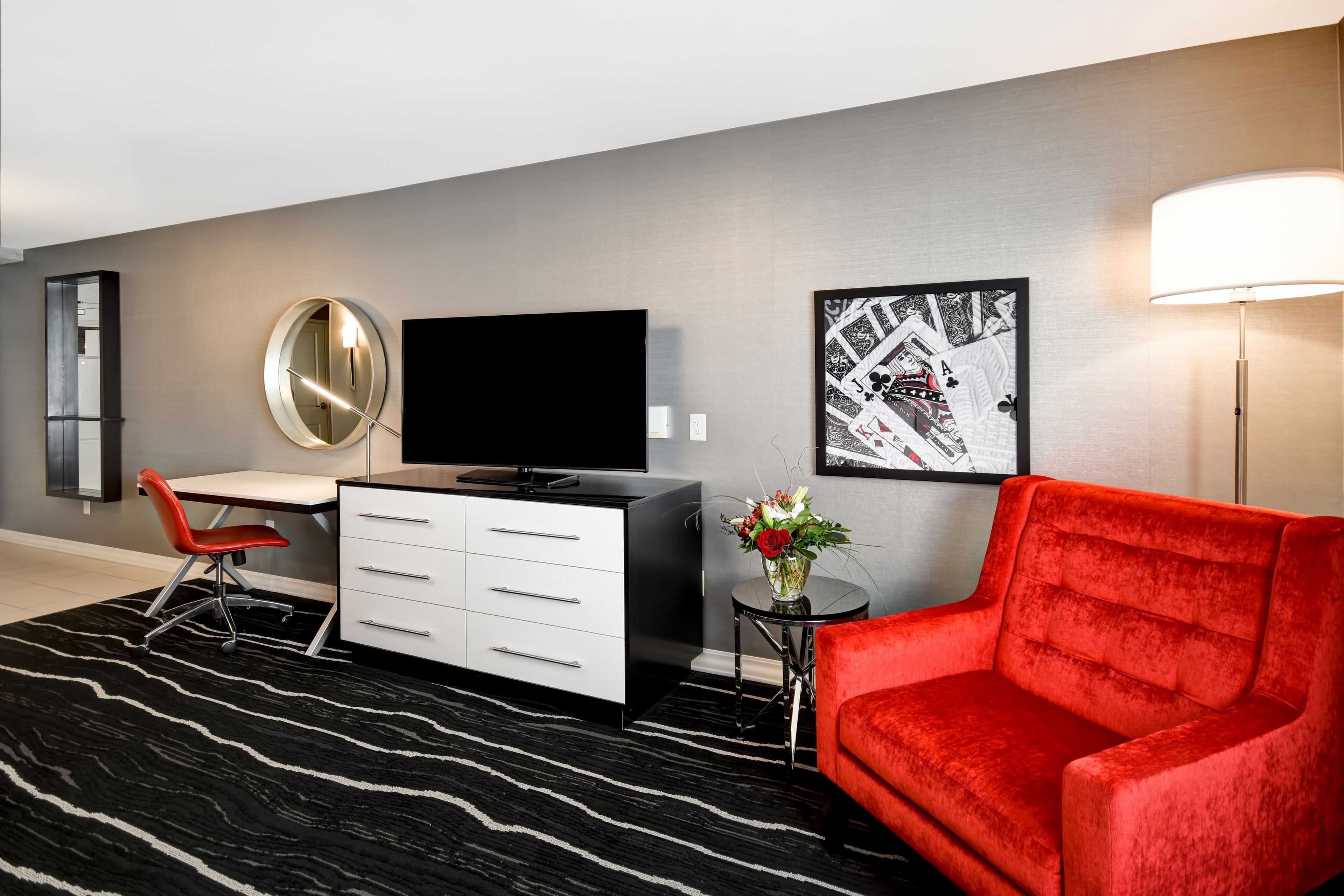 A modern hotel room with a black and white striped carpet, a red sofa, a white desk with drawers, a flat-screen TV, a round mirror, a small side table with a vase of flowers, and two framed artworks on the wall.
