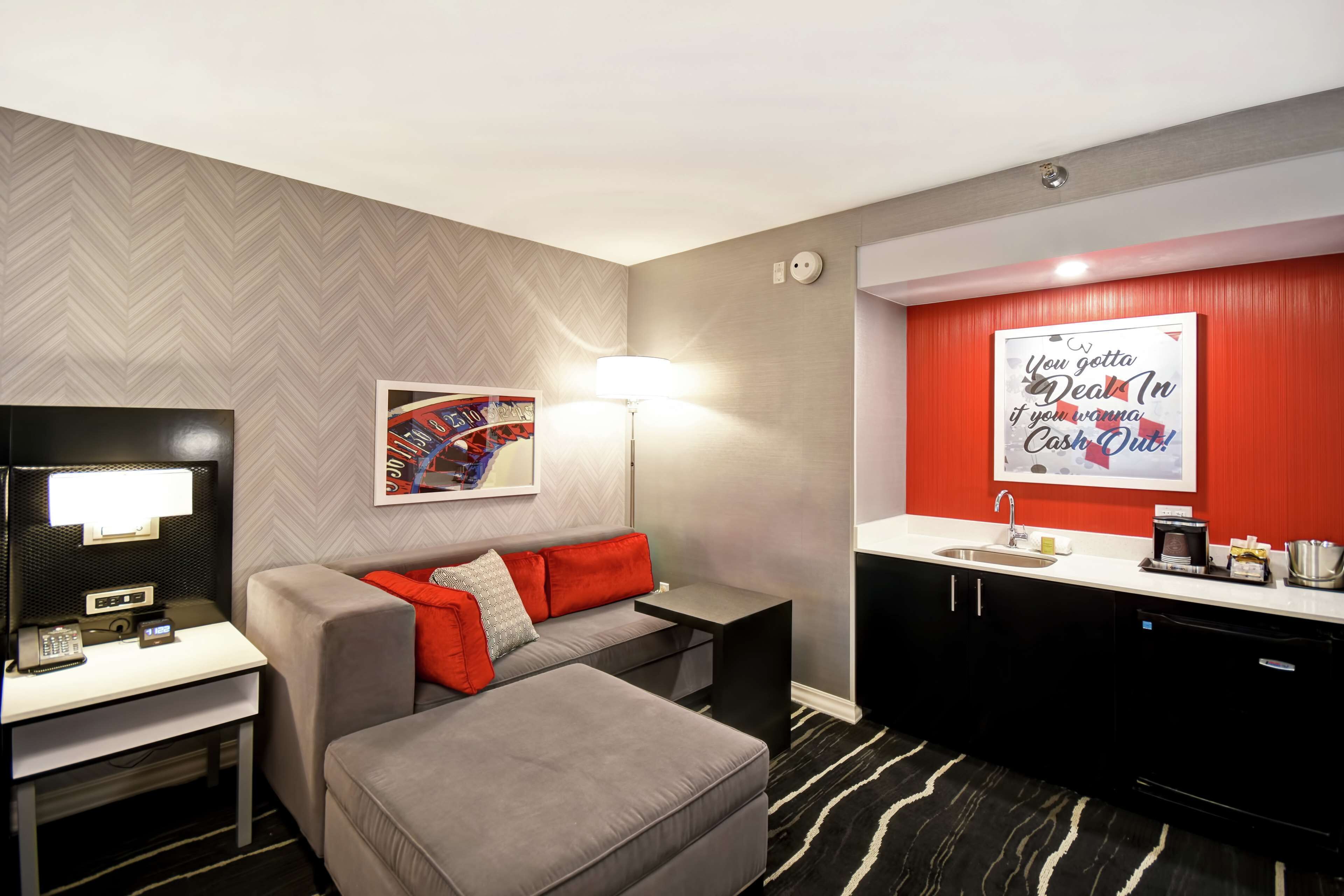 Una habitación de hotel moderna con un sofá gris, un escritorio pequeño con lámpara, una pared de acento roja y una zona de cocina con fregadero y cafetera.