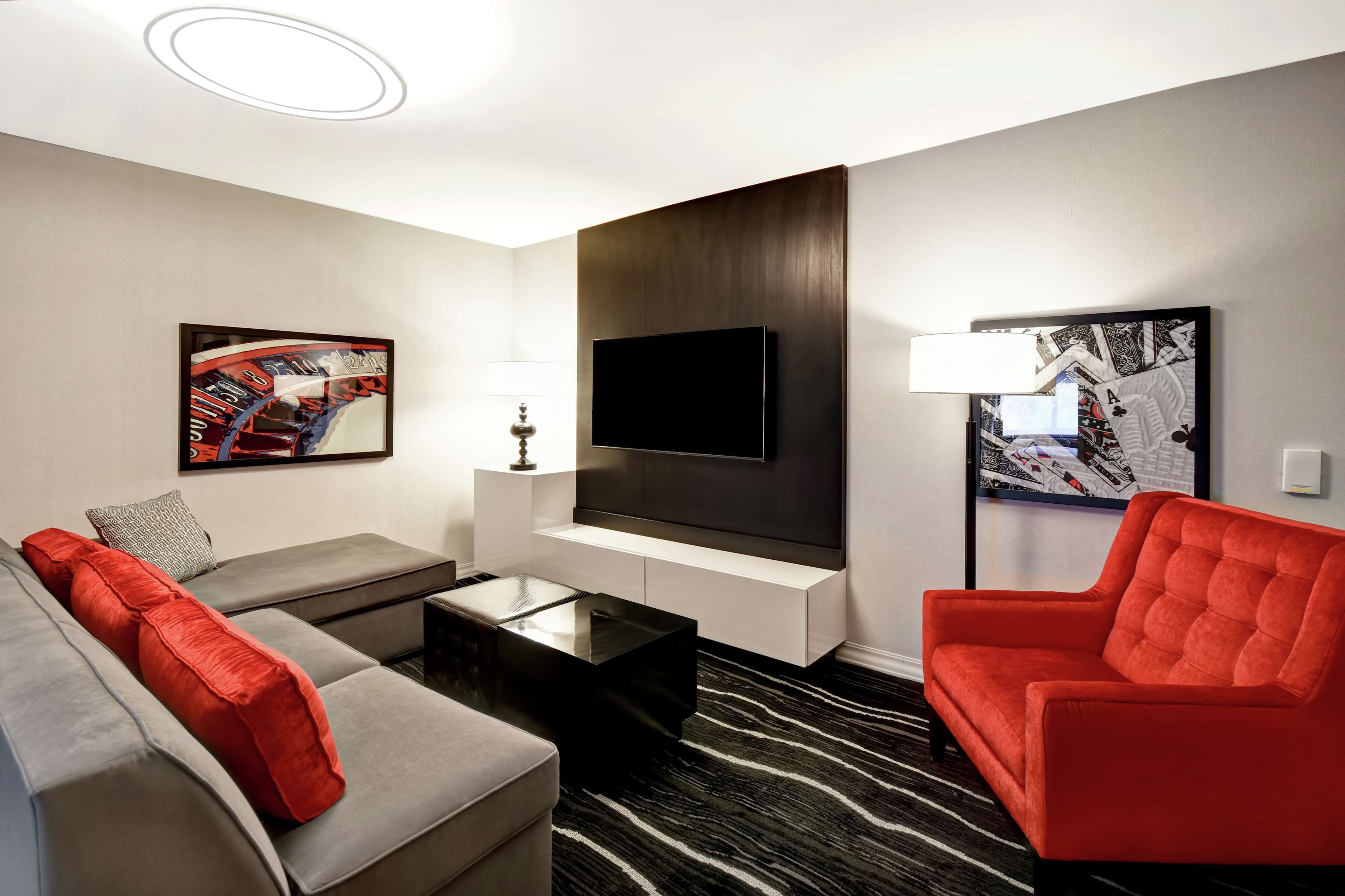 A modern hotel room with a grey sofa, red armchair, and a flat-screen TV mounted on the wall.