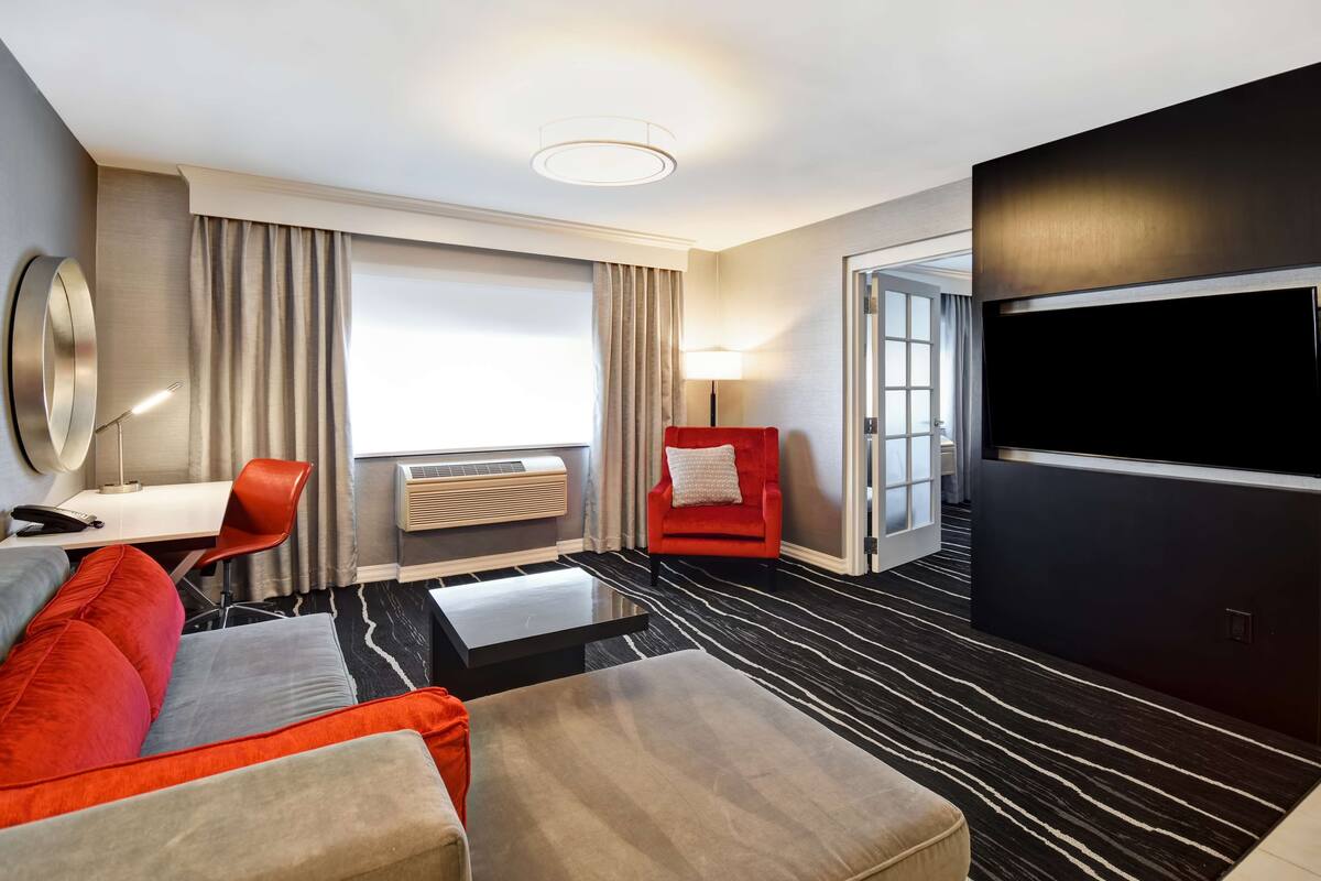 Habitación de hotel con diseño moderno, que cuenta con un televisor de pantalla plana grande, un sofá, un sillón rojo, un escritorio con lámpara y un amplio ventanal con cortinas.