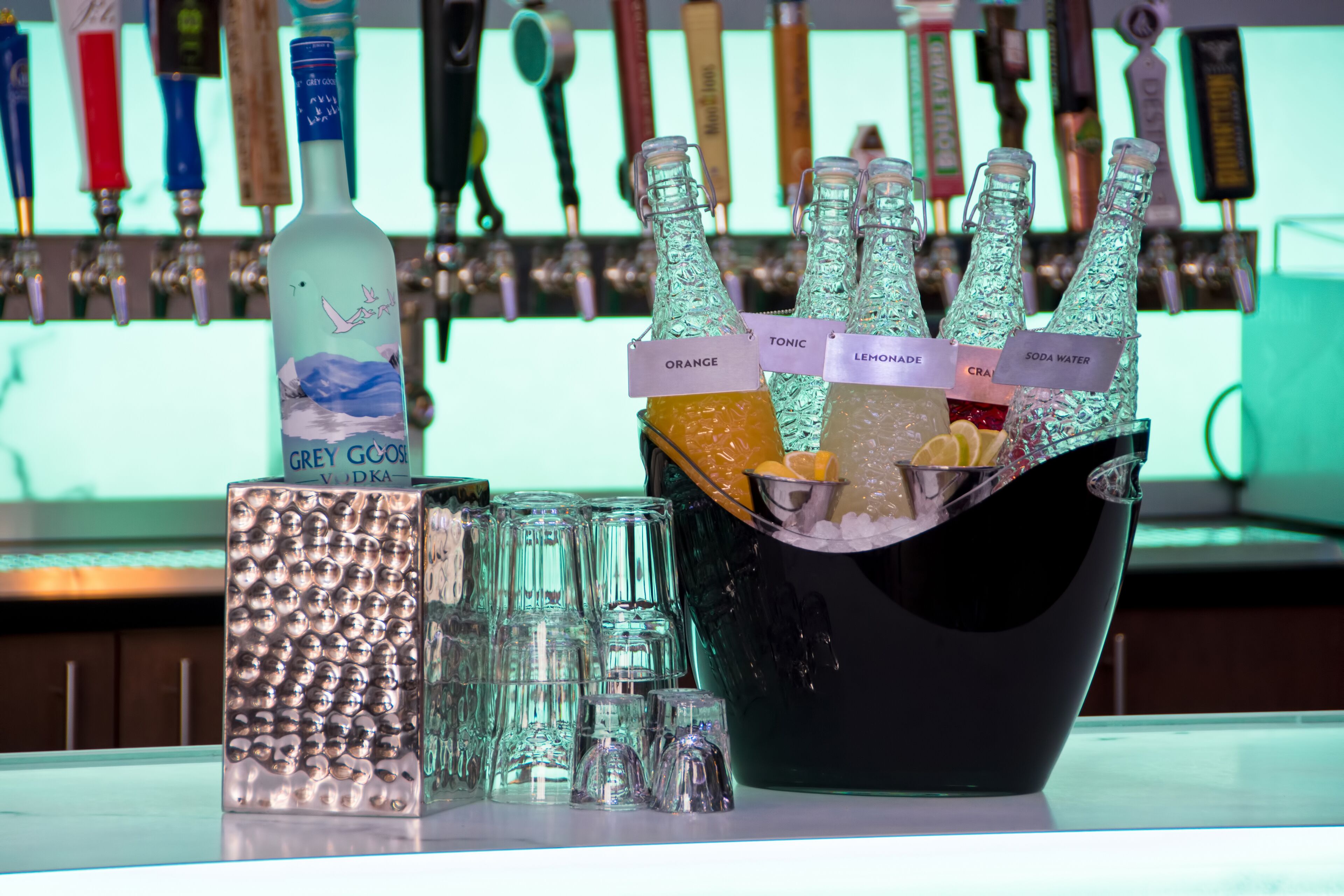 A bar area with a bottle of Grey Goose vodka, a bucket of ice with various bottles labeled with different types of beverages, and several glasses.