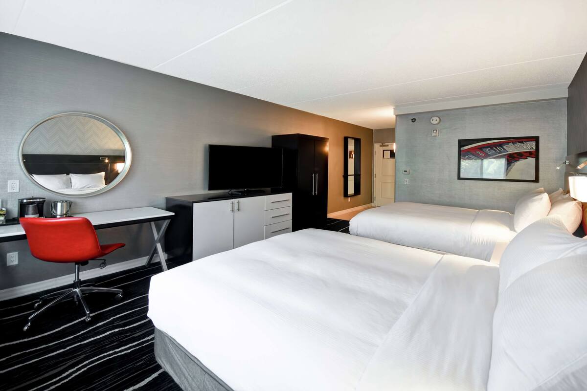 Una habitación de hotel con dos camas, un escritorio, una silla roja, un televisor y un espejo.