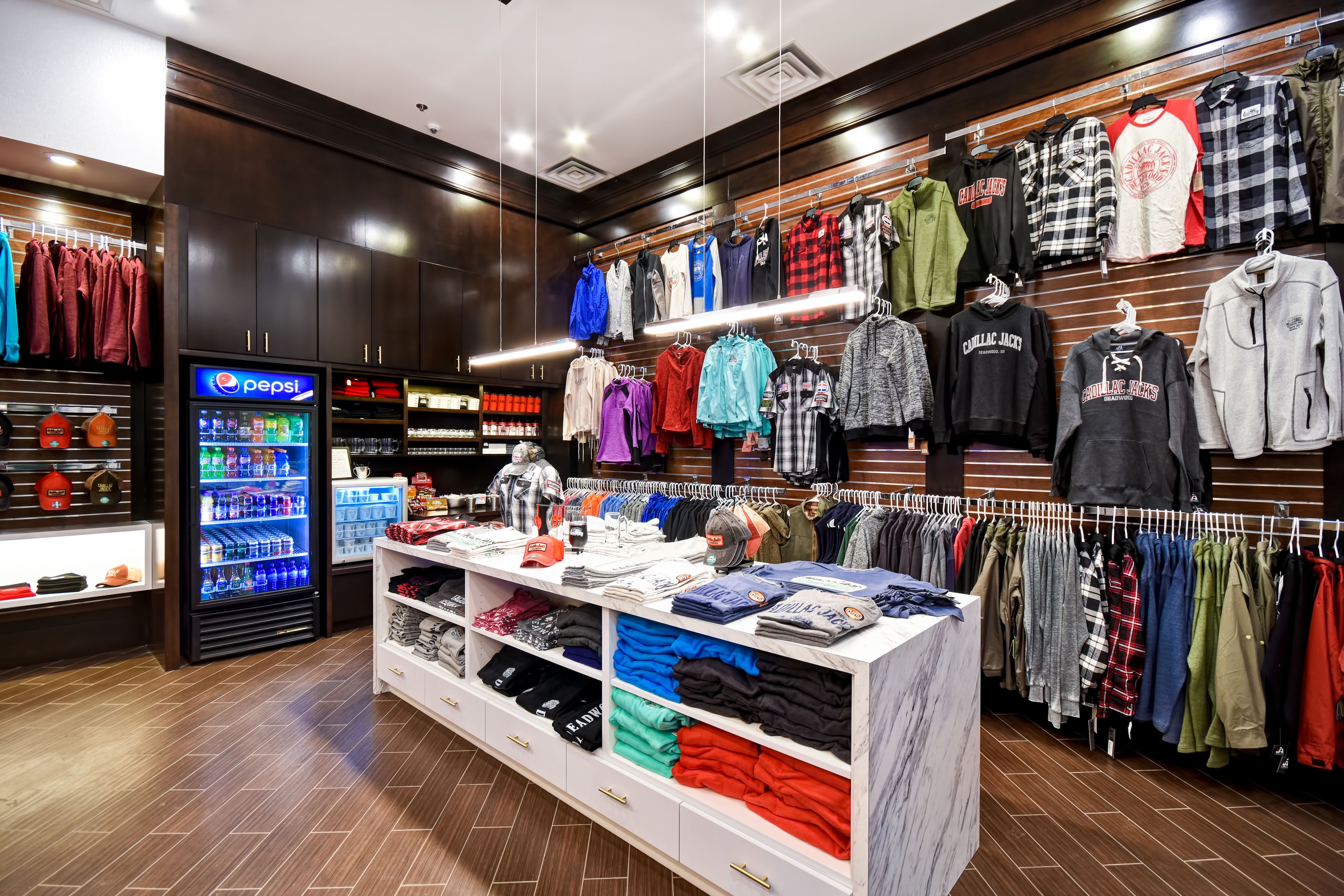 A clothing store with racks of shirts and jackets, a Pepsi refrigerator, and a display of folded clothes.