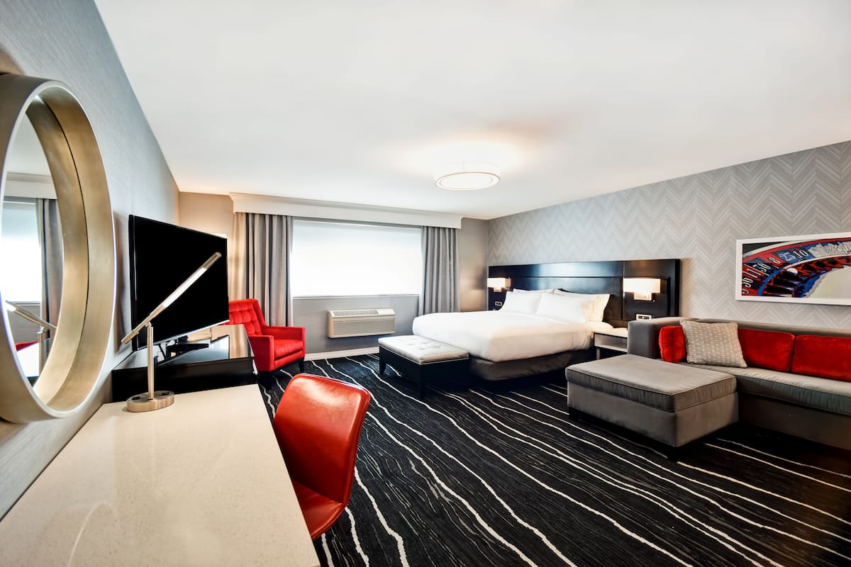 Habitación de hotel con cama, escritorio, silla, sofá, televisor y un espejo grande.