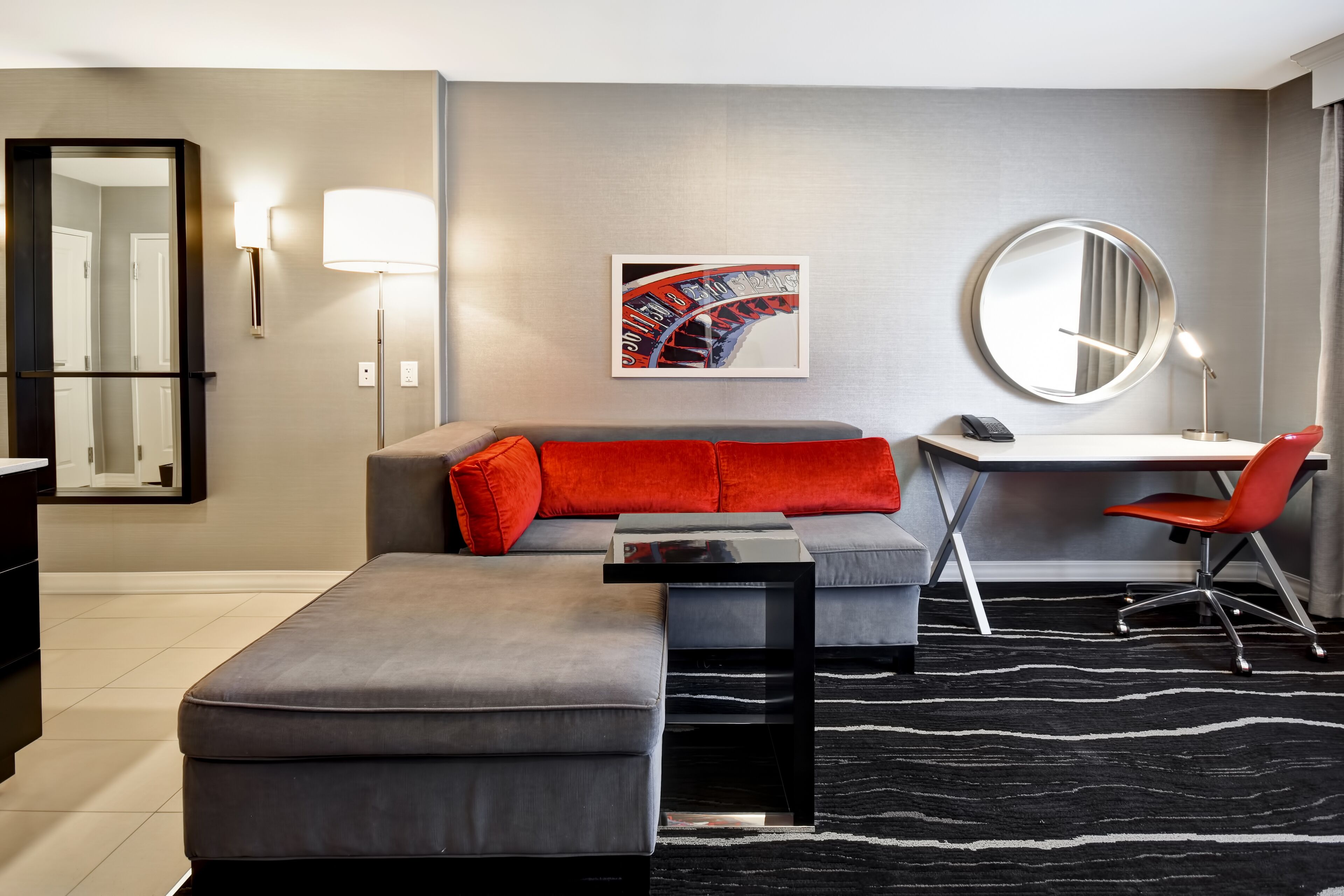 A modern hotel room with a grey sectional sofa, a small desk, a round mirror, and a black and white patterned rug.
