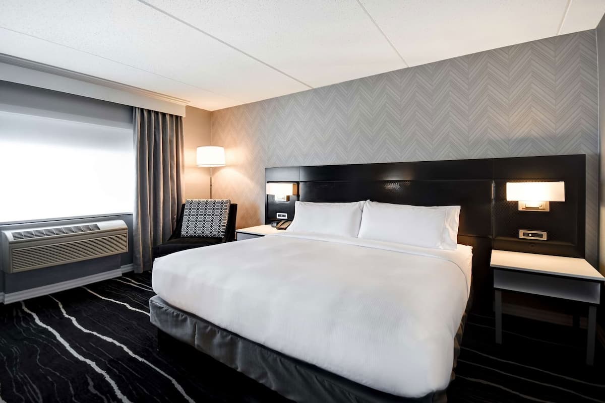 A hotel room with a large bed, a desk, a chair, a nightstand, a lamp, and a window with curtains.