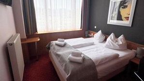 Habitación de hotel con cama, dos almohadas, mesita de noche, radiador y ventana con cortinas.