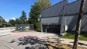 Un estacionamiento vallado con entrada a un edificio, un camino peatonal y vehículos estacionados.