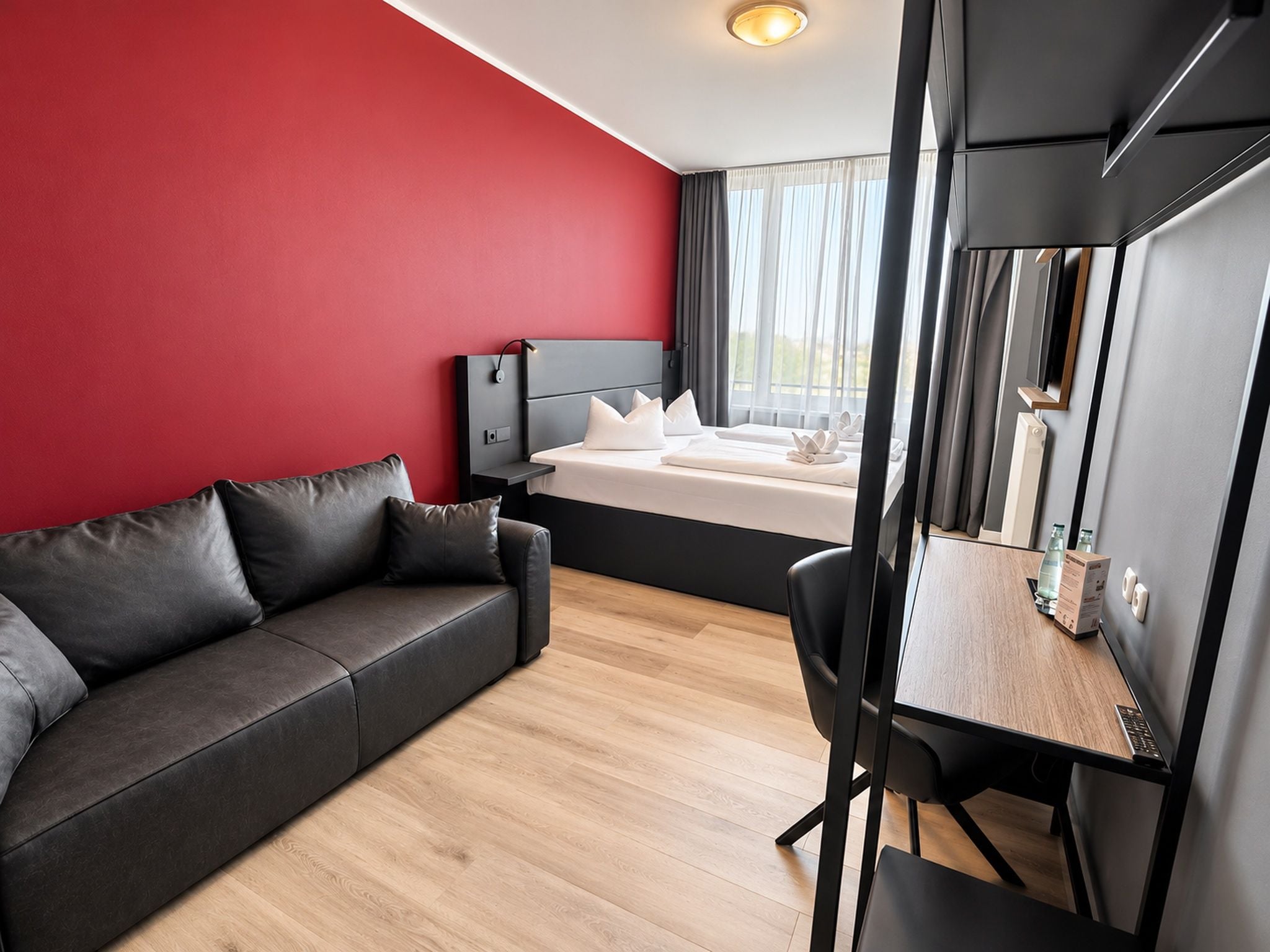 Ein modernes Hotelzimmer mit roter Akzentwand, einem grauen Sofa, einem Bett mit weißen Leinentüchern, einem Schreibtisch mit Computer und einem großen Spiegel.