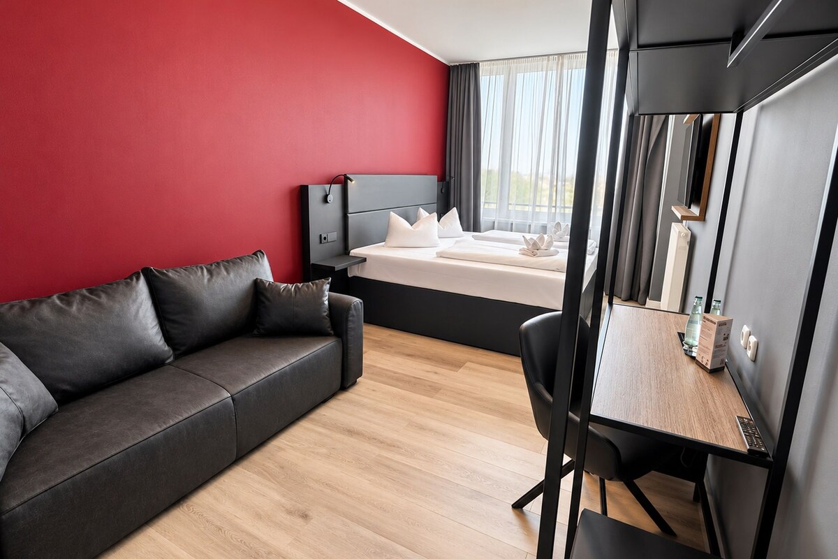 A modern hotel room with a red accent wall, a grey sofa, a bed with white linens, a desk with a computer, and a large mirror.