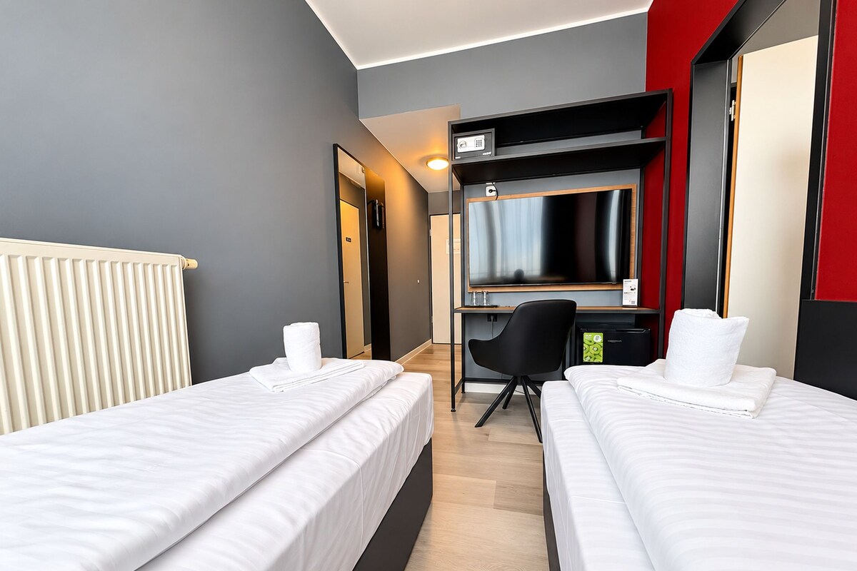 A hotel room with two beds, a desk, a chair, a TV, and a radiator.