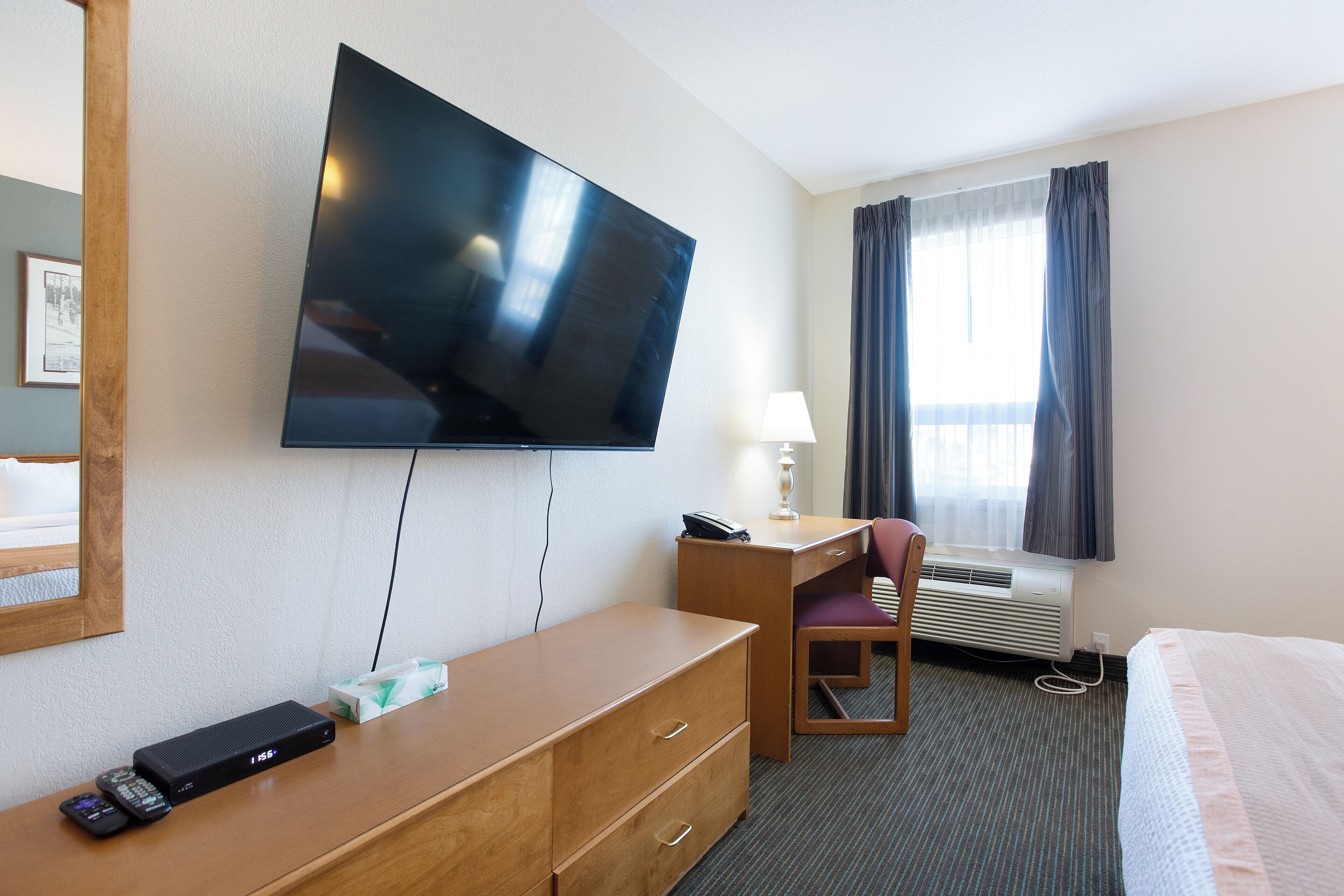 A hotel room with a flat-screen TV mounted on the wall, a desk with a chair, a bed, a bedside table, and a window with curtains.