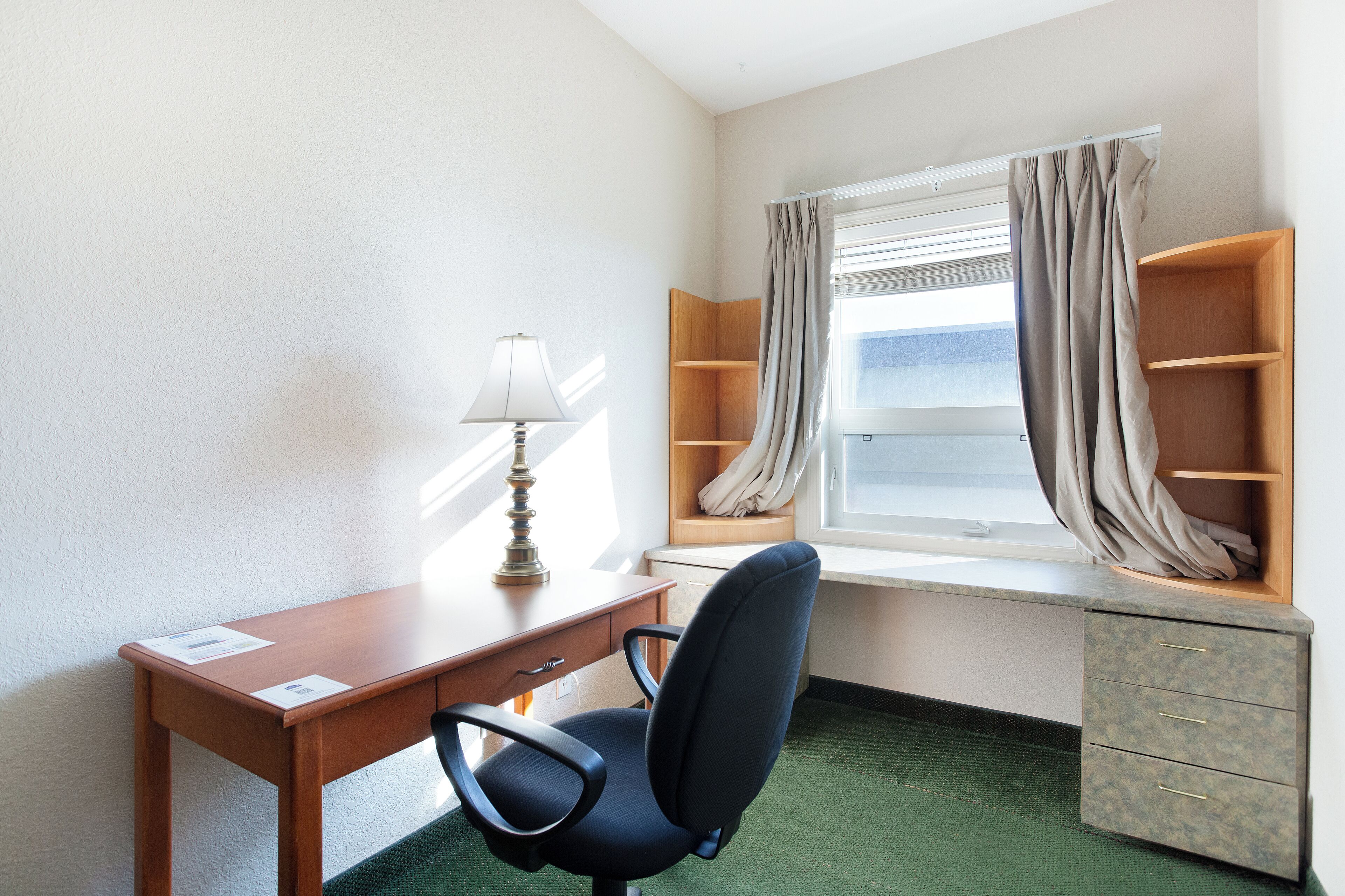 Habitació petita i ben ordenada amb escriptori, cadira, làmpada, finestra amb cortines i prestatge incorporat.