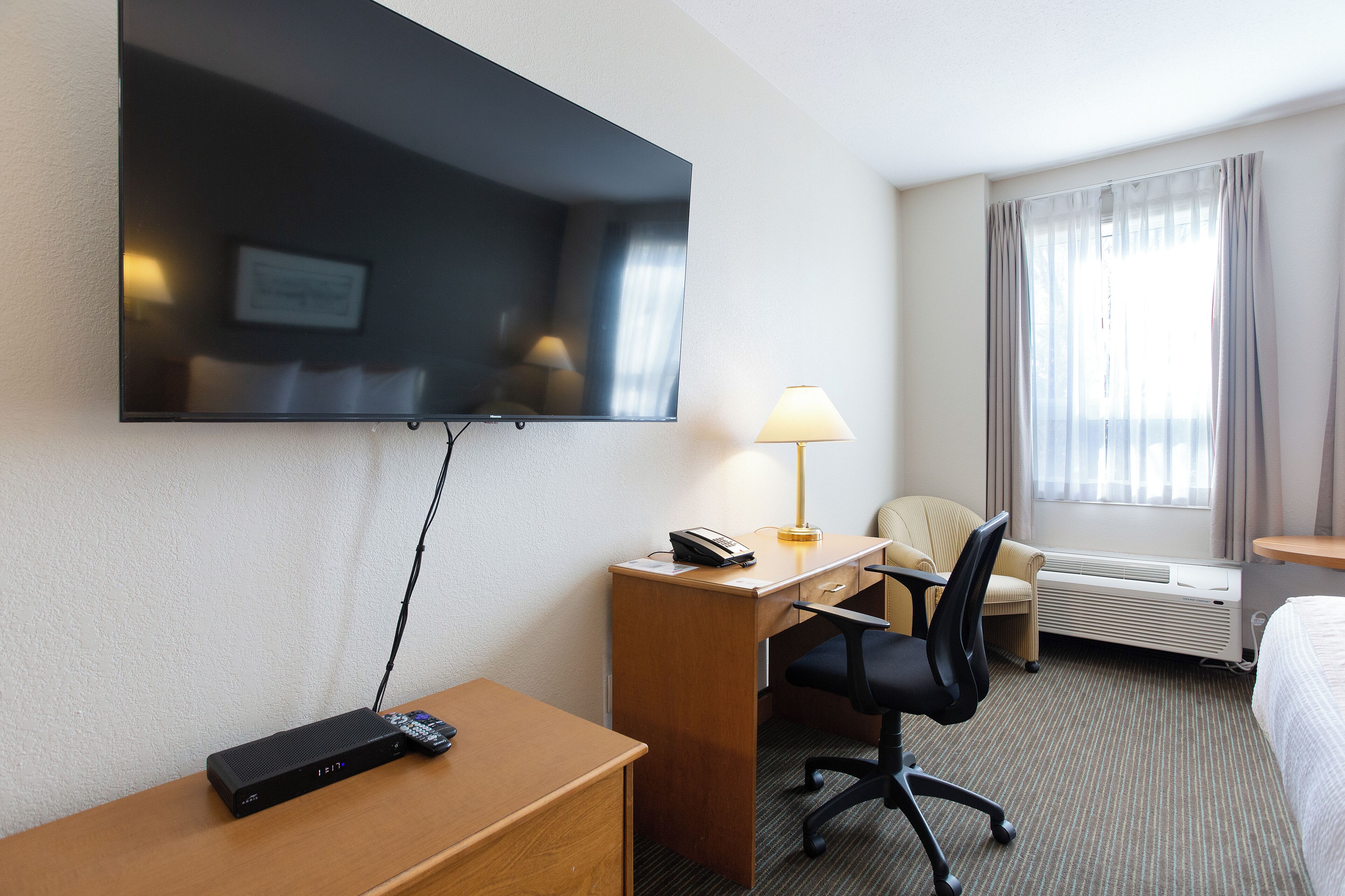 Een hotelkamer met een flatscreen televisie aan de muur, een bureau met een lamp, een stoel en een bed.