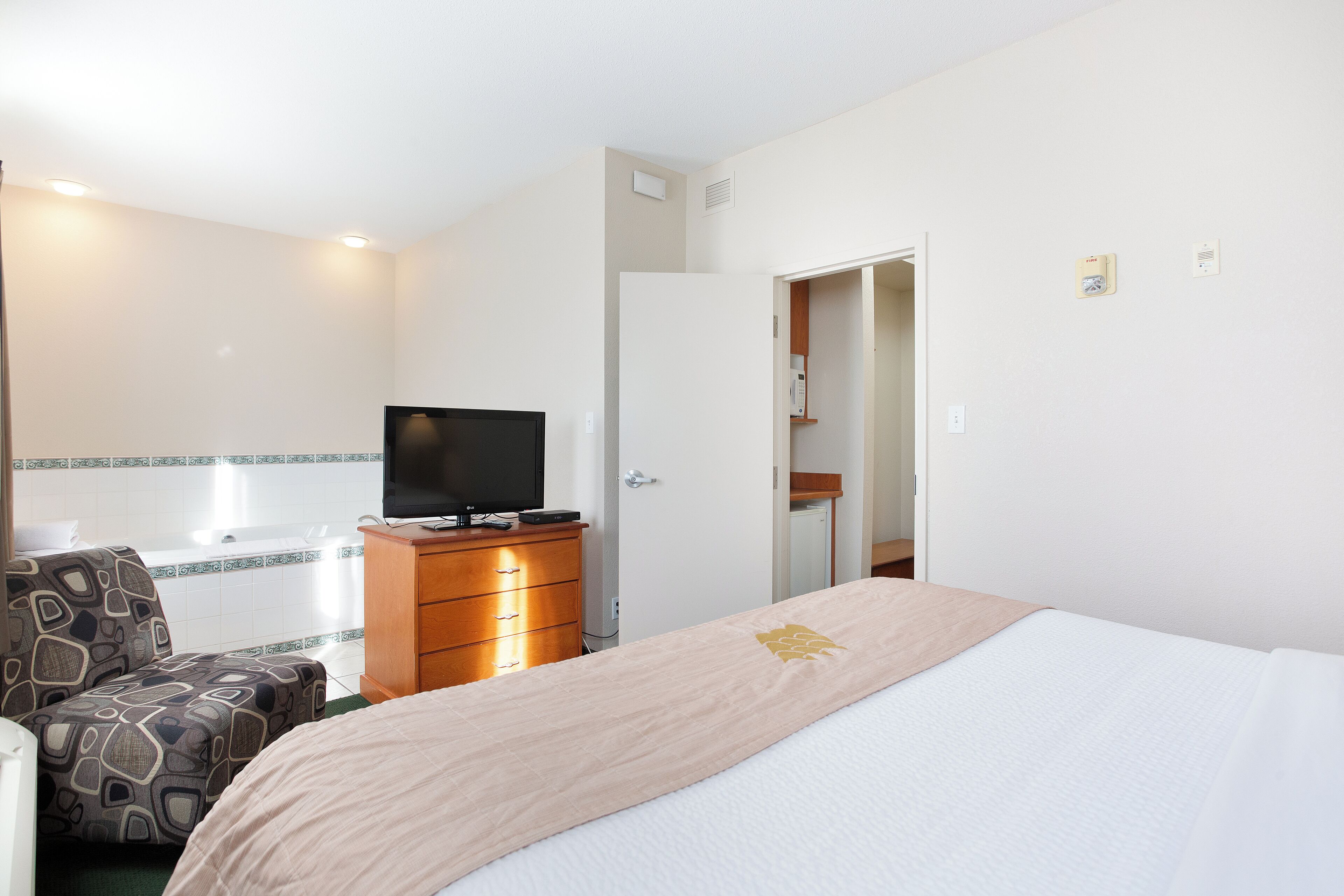 A hotel room with a bed, a TV on a wooden dresser, a patterned armchair, and a bathroom visible through an open door.