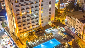 A high-rise hotel with a rooftop pool area, multiple balconies, and a view of the cityscape at night.