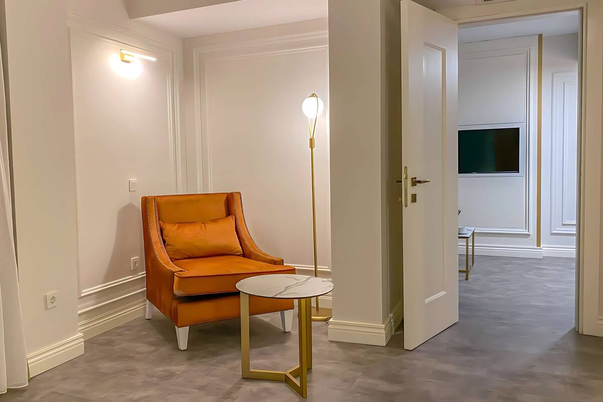 Een kamer met een losse oranje fauteuil, een klein rond tafeltje, een vloerlamp en een deur naar een andere kamer.