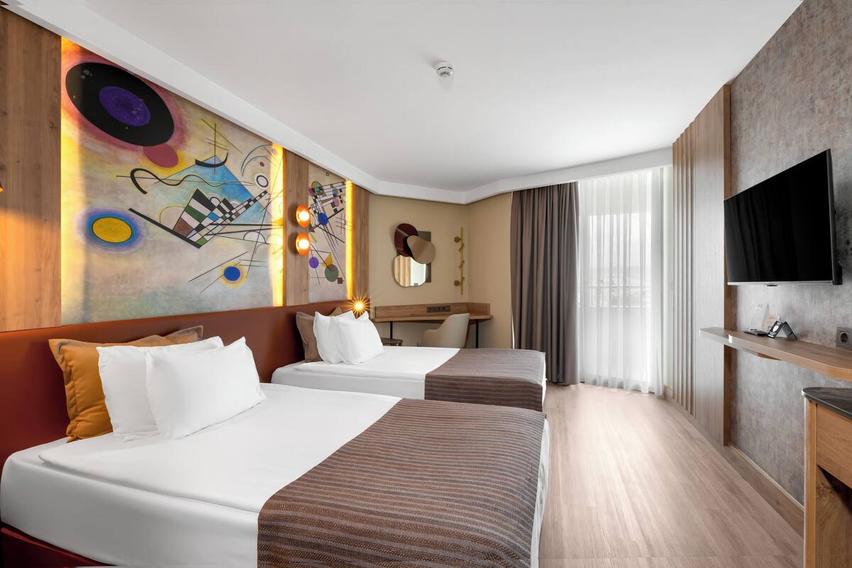 A hotel room with two beds, a desk, a TV, and a large abstract painting on the wall.