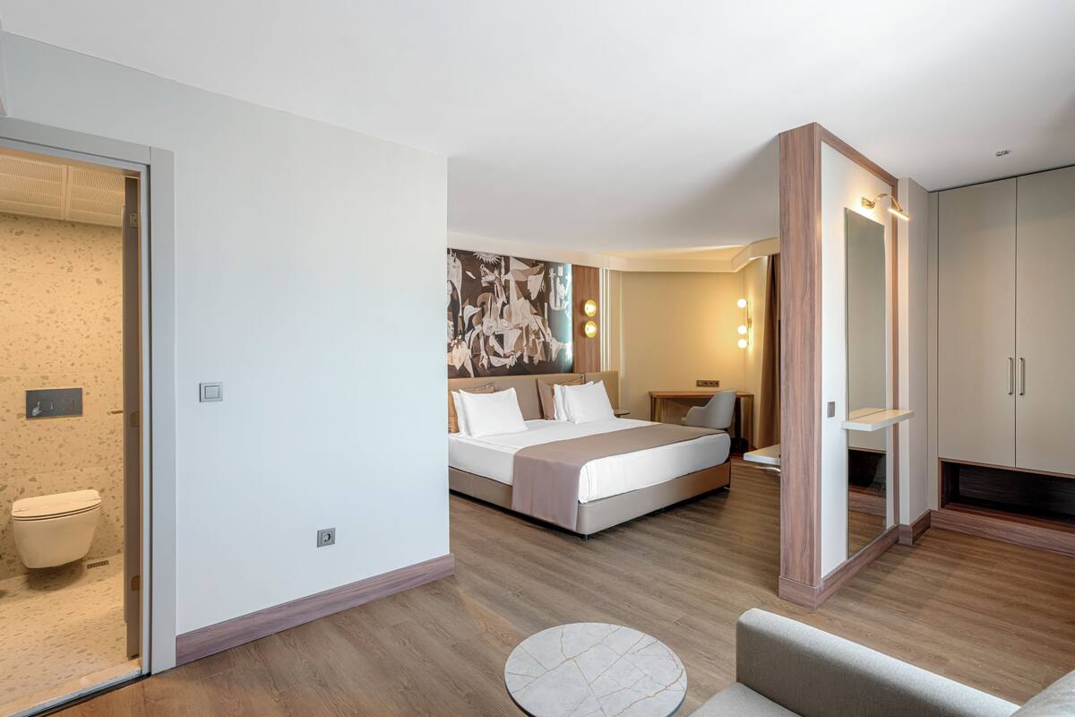 A modern hotel room with a bed, a round coffee table, a bathroom with a toilet, and a large mirror.