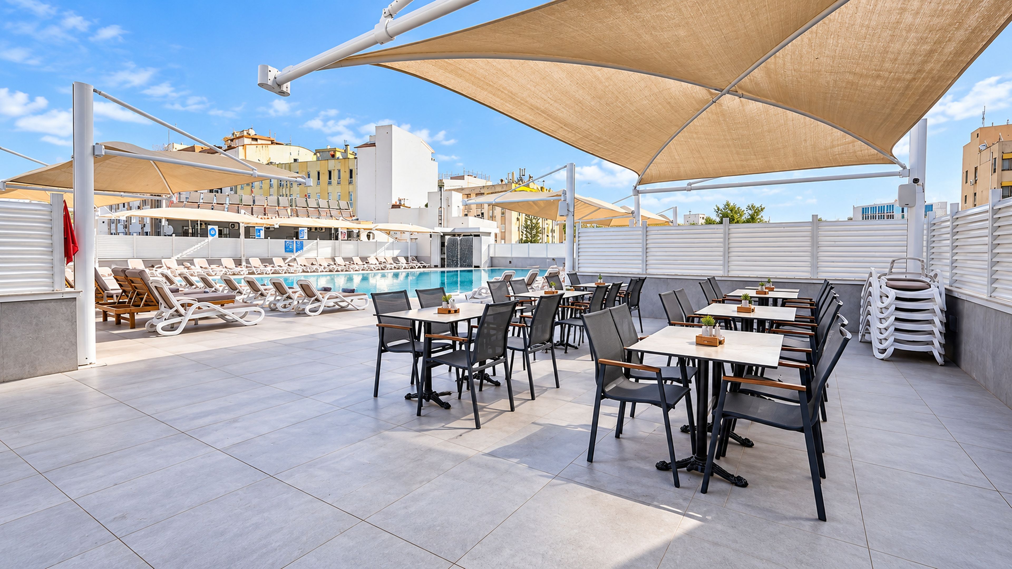 A rooftop pool area with sun loungers, tables, and chairs, surrounded by buildings.