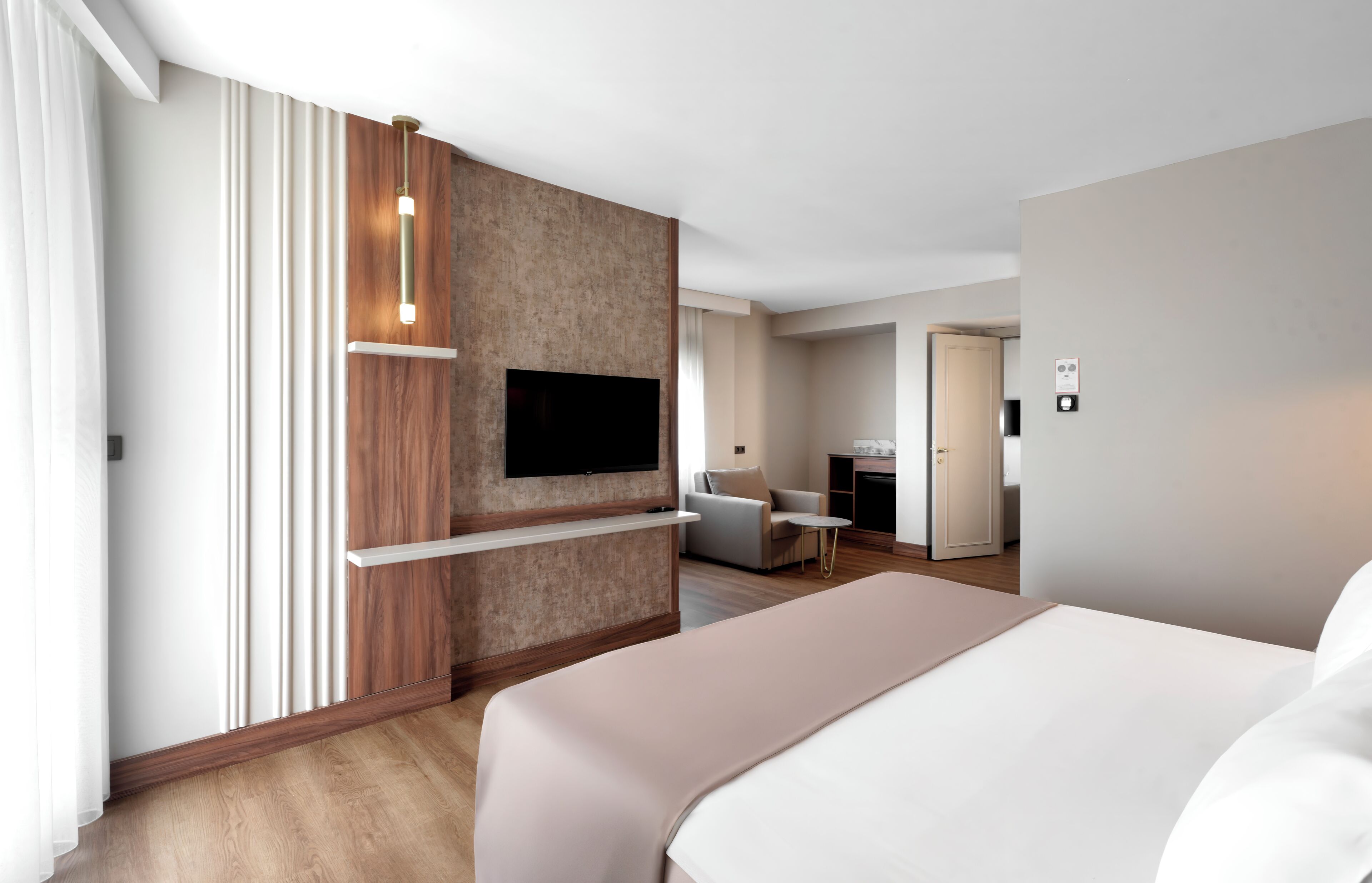 Ein modernes Hotelzimmer mit einem großen Bett, einem Flachbildfernseher an einer Holzvertäfelung, einem kleinen runden Tisch und einer separaten Sitzecke mit Sofa und Sessel.