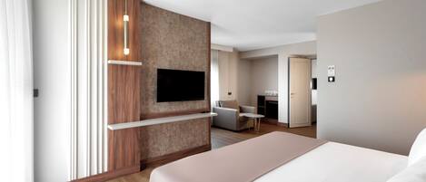 A modern hotel room with a large bed, a flat-screen TV mounted on a wooden panel, a small round table, and a separate seating area with a sofa and chair.