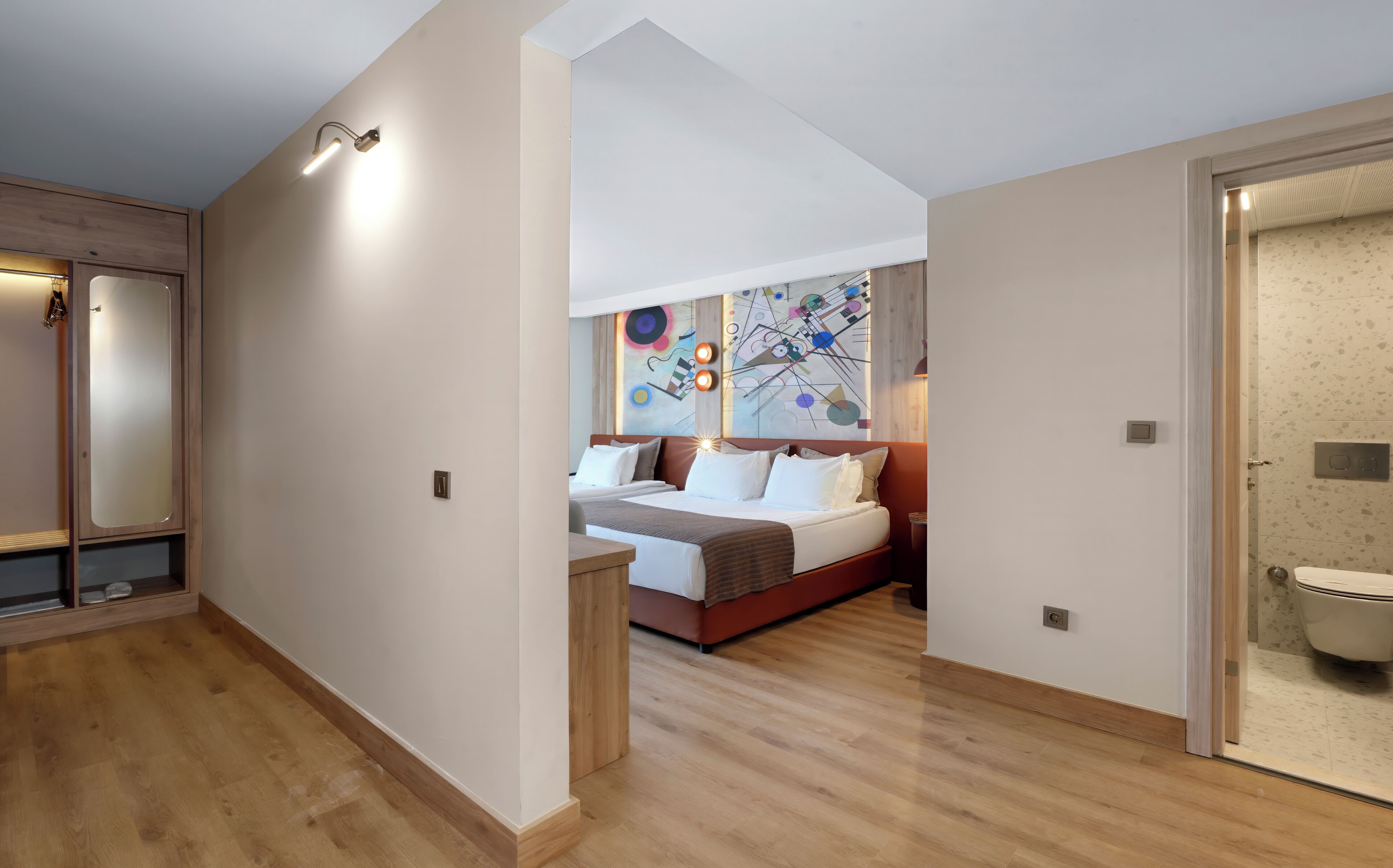 A modern hotel room with a wooden floor, a bed with white bedding, a colorful abstract wall art, and a bathroom with a toilet and sink.