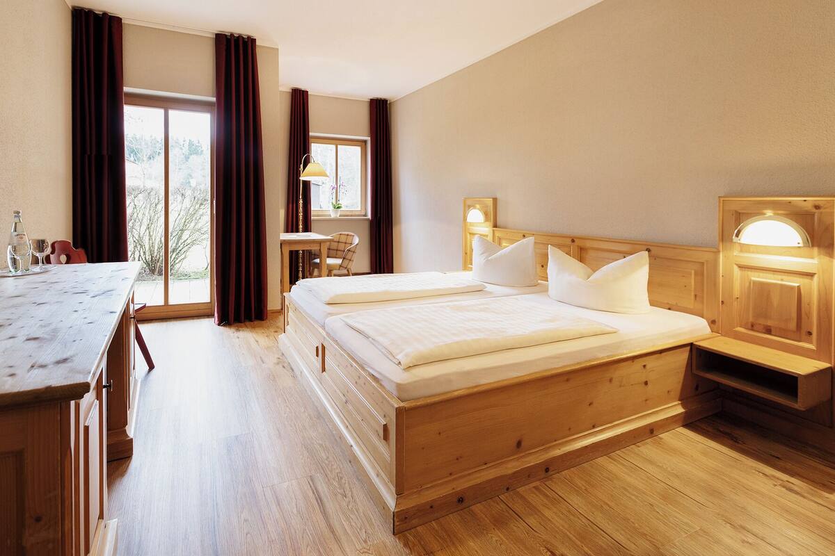 A hotel room with a wooden bed, a desk, a chair, a small table, and a window with curtains.
