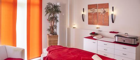 A modern spa room with a massage table, a white chair, a plant, a sink, and a red towel.