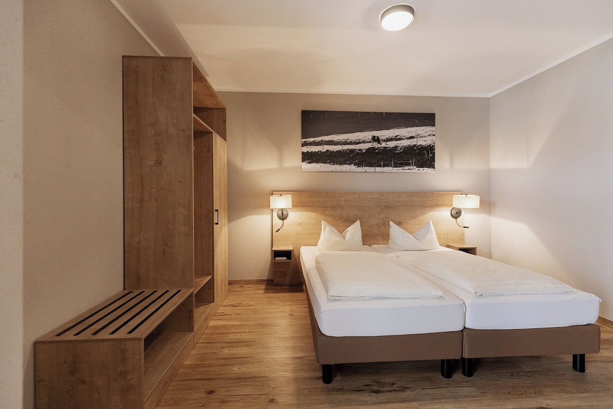 A hotel room with a wooden wardrobe, a bed with white linens, two bedside lamps, and a wall-mounted picture.