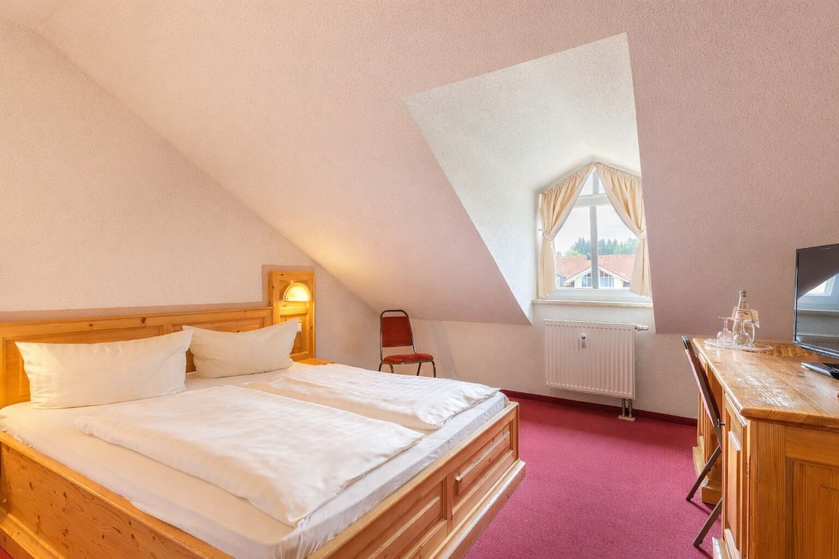 A bedroom with a wooden bed, a chair, a desk with a computer, a window with curtains, and a radiator.