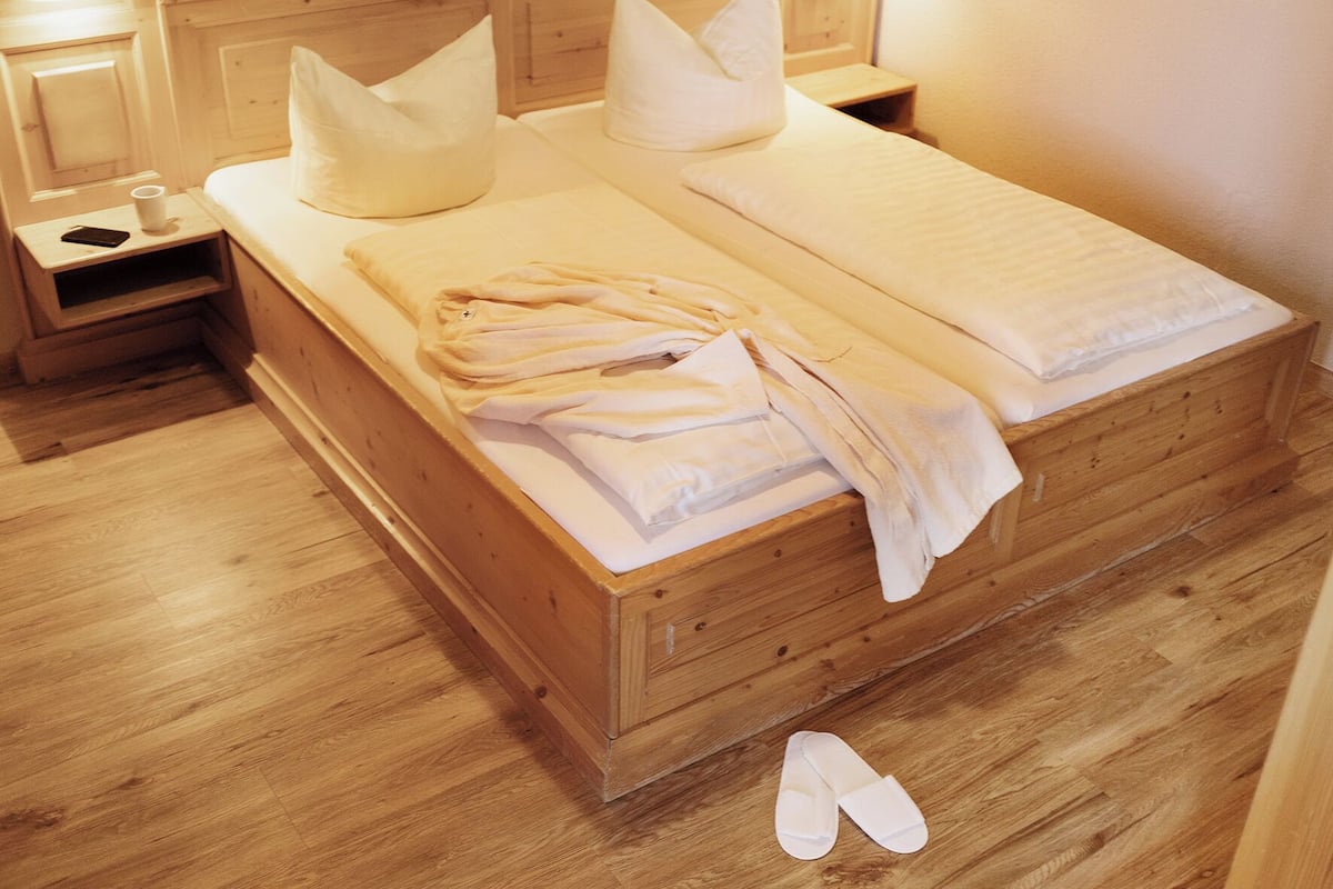 A wooden bed with white linens and pillows, a bedside table with a cup and a book, and a pair of slippers on the floor.
