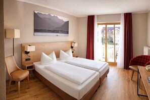 A hotel room with two beds, a wooden headboard, a painting of mountains, a desk with a chair, and a view of buildings through a window. - Hotel Ahornhof (Lindberg)