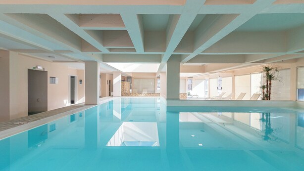 A large indoor swimming pool with clear water, surrounded by walls and columns, and a few lounge chairs by the pool. - Alvor Atlântico Residences (Portimão)