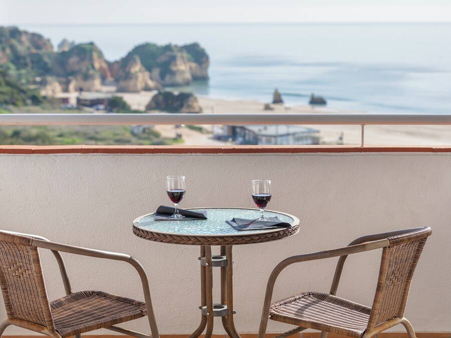 Ein Balkon mit zwei Korbstühlen und einem kleinen Tisch, mit Blick auf einen Strand mit steinigen Klippen und dem Meer.