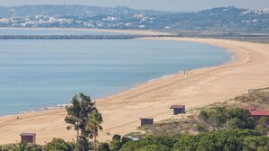 A coastal beach with a rocky breakwater, sandy shore, and a few buildings in the background. - Alvor Atlântico Residences (Portimão)