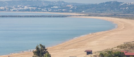 A coastal beach with a rocky breakwater, sandy shore, and a few buildings in the background.