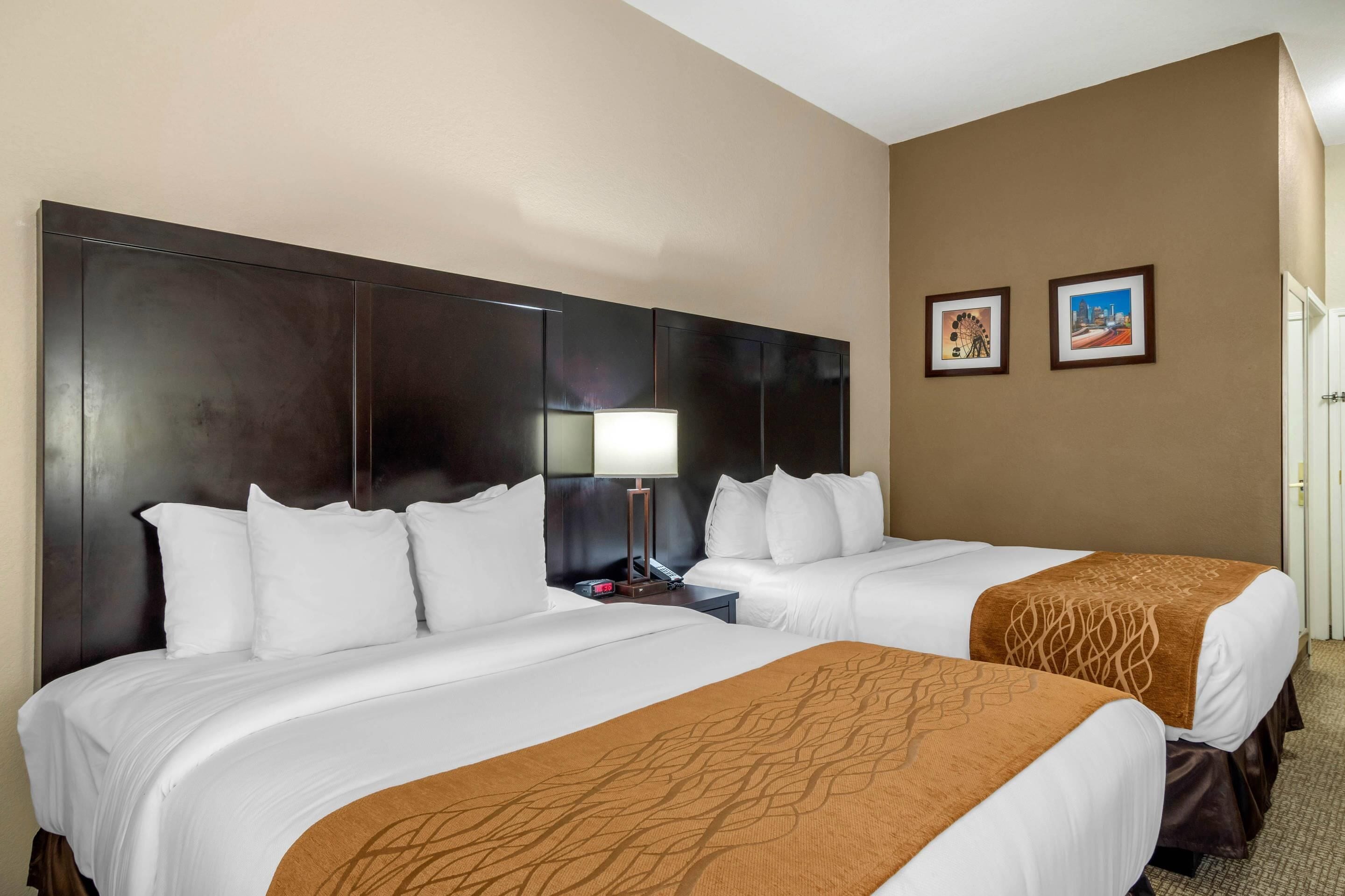 A hotel room with two beds, a large dark headboard, a nightstand with a lamp, and two framed pictures on the wall.